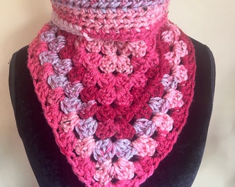 May include: A handmade crochet cowl in shades of pink, red, and purple. The cowl features a granny square design and a ribbed turtleneck. The yarn appears to be a soft, bulky weight, perfect for keeping warm.