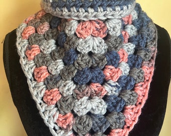 May include: A handmade crochet cowl scarf in a granny square pattern. The scarf features a mix of grey, navy blue, and peach-coloured yarn. The cowl is displayed on a black mannequin head against a yellow background.