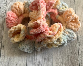 May include: A collection of pastel-colored scrunchies. The scrunchies are made of a soft, plush material in shades of pink, yellow, and blue. The scrunchies are arranged in a circular shape, with the fabric bunched up to create a textured look. Hair accessories.