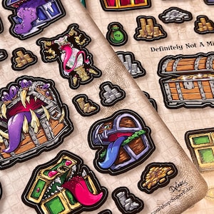 May include: A sheet of fantasy-themed stickers featuring treasure chests with sharp teeth and long tongues. The stickers are in various colors, including brown, gold, purple, and green. The text "Definitely Not A Mimic" is visible.