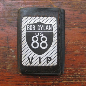 May include: Black leather wallet with a Bob Dylan US 88 VIP patch. The patch is black and white with the text "BOB DYLAN", "US", "88", and "VIP". The wallet is closed and appears worn.