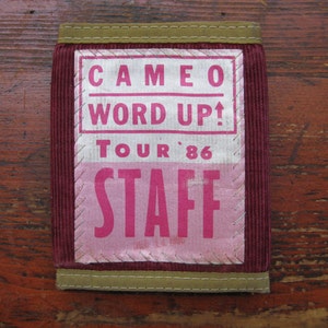 May include: A burgundy corduroy wallet with a rectangular patch featuring the text "CAMEO WORD UP! TOUR '86 STAFF" in pink and white. The wallet has a khaki-colored trim.