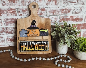 Może przedstawiać: Drewniany znak Halloweenowy z dynią, czarodziejskim kapeluszem i kotłem. Znak głosi "HALLOWEEN" czarnymi literami na tle tablicy.