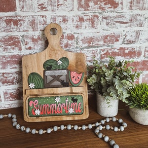 May include: Wooden cutting board with a green and red watermelon, a box labeled "Watermelon", and a green and white floral design. The board has a shelf with the word "Summertime" written in green and red letters.