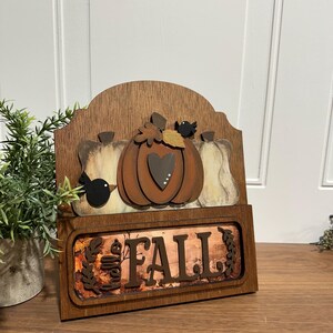 May include: Wooden sign with a autumn theme. It features three pumpkins, two birds, and the word "FALL" in brown letters on a background of autumn leaves.