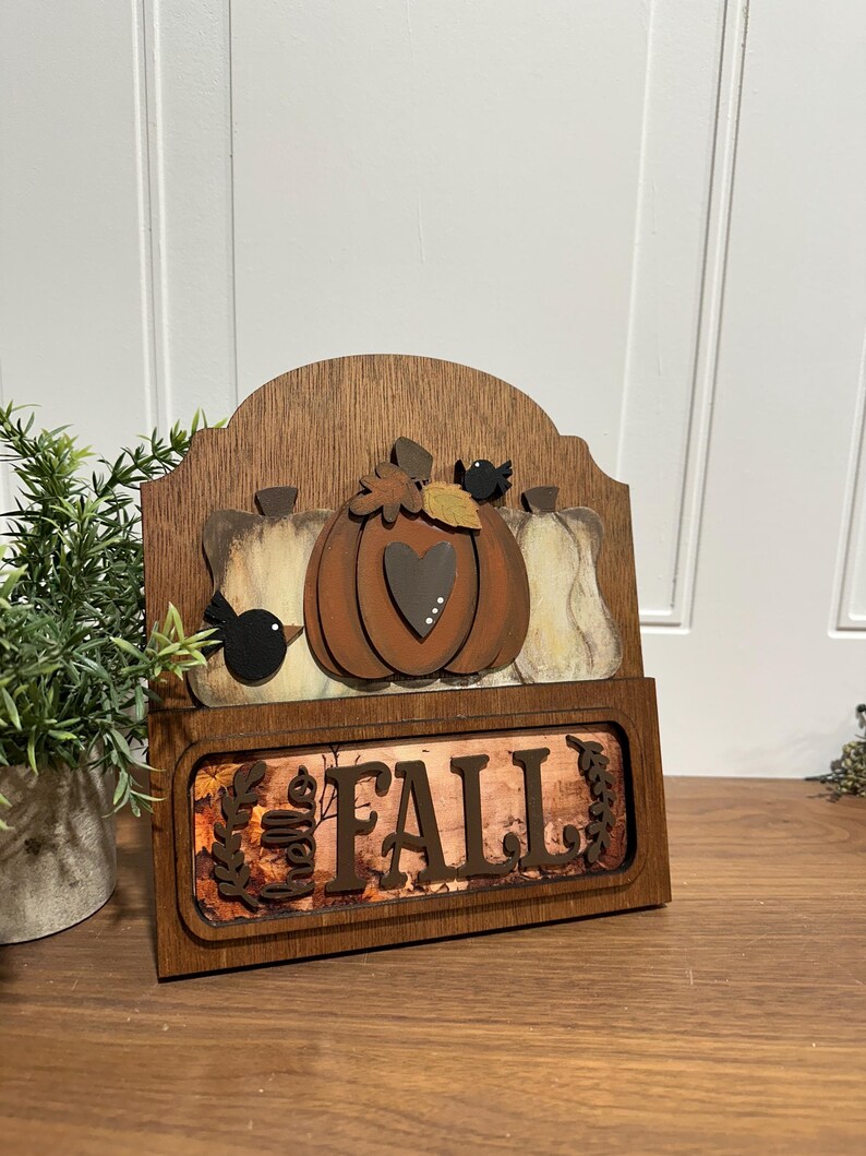 May include: Wooden sign with a autumn theme. It features three pumpkins, two birds, and the word "FALL" in brown letters on a background of autumn leaves.