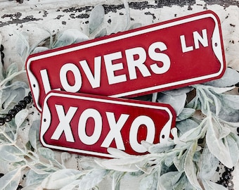 May include: Two red wooden signs with white lettering. The top sign reads "LOVERS LN" and the bottom sign reads "XOXO".