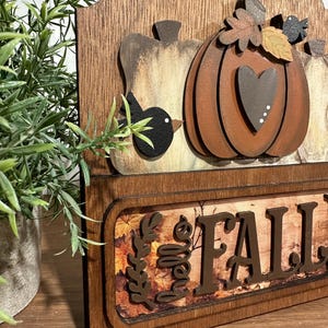 May include: Wooden sign featuring a carved pumpkin, leaves, and birds. The sign reads "Hello Fall" in brown lettering against a backdrop of autumn leaves.