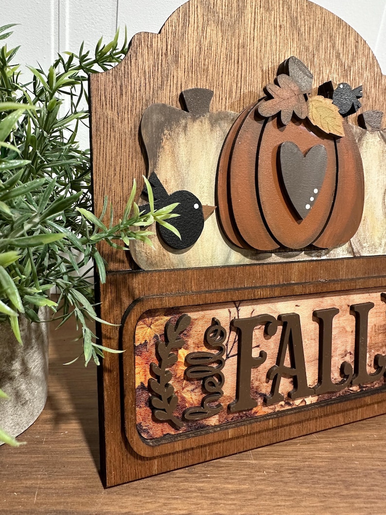 May include: Wooden sign featuring a carved pumpkin, leaves, and birds. The sign reads "Hello Fall" in brown lettering against a backdrop of autumn leaves.