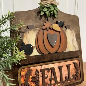 May include: A wooden chopping board with a autumnal theme. It features a pumpkin with a heart, leaves, and birds. The bottom of the board has a sign that says "Fall" with a brown background and leaves.