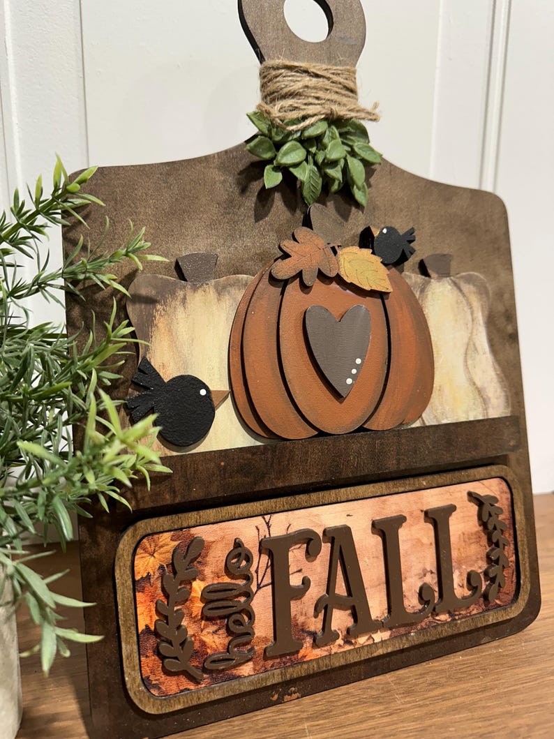 May include: A wooden chopping board with a autumnal theme. It features a pumpkin with a heart, leaves, and birds. The bottom of the board has a sign that says "Fall" with a brown background and leaves.