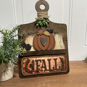 May include: A wooden chopping board with a autumnal theme. It features a pumpkin with a heart, two birds, and the word "FALL" on a copper-coloured background.