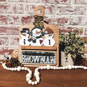 May include: Wooden sign with three snowmen and the text "In the window we can build a SNOWMAN". The snowmen are painted white with black eyes and buttons, and one has a red heart. The sign is decorated with green leaves and a brown twine bow.