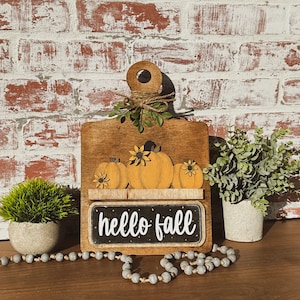 May include: Wooden cutting board with a chalkboard sign that says "hello fall". The board is decorated with three painted pumpkins and fall leaves.