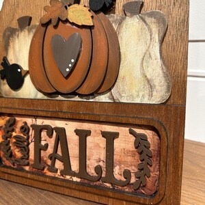 May include: Wooden autumn sign featuring a brown pumpkin, gourds, and birds. The sign reads "Hello Fall" with brown leaves on a woodgrain background.
