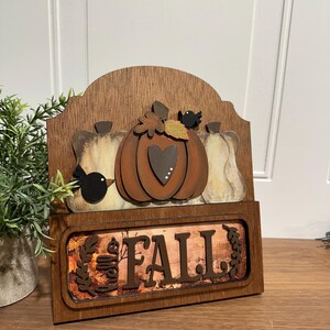 May include: Wooden sign with a autumnal theme. It features a brown pumpkin with a heart, two white pumpkins, and two black birds. The sign also has a brown background with the words "Hello Fall" in white letters.