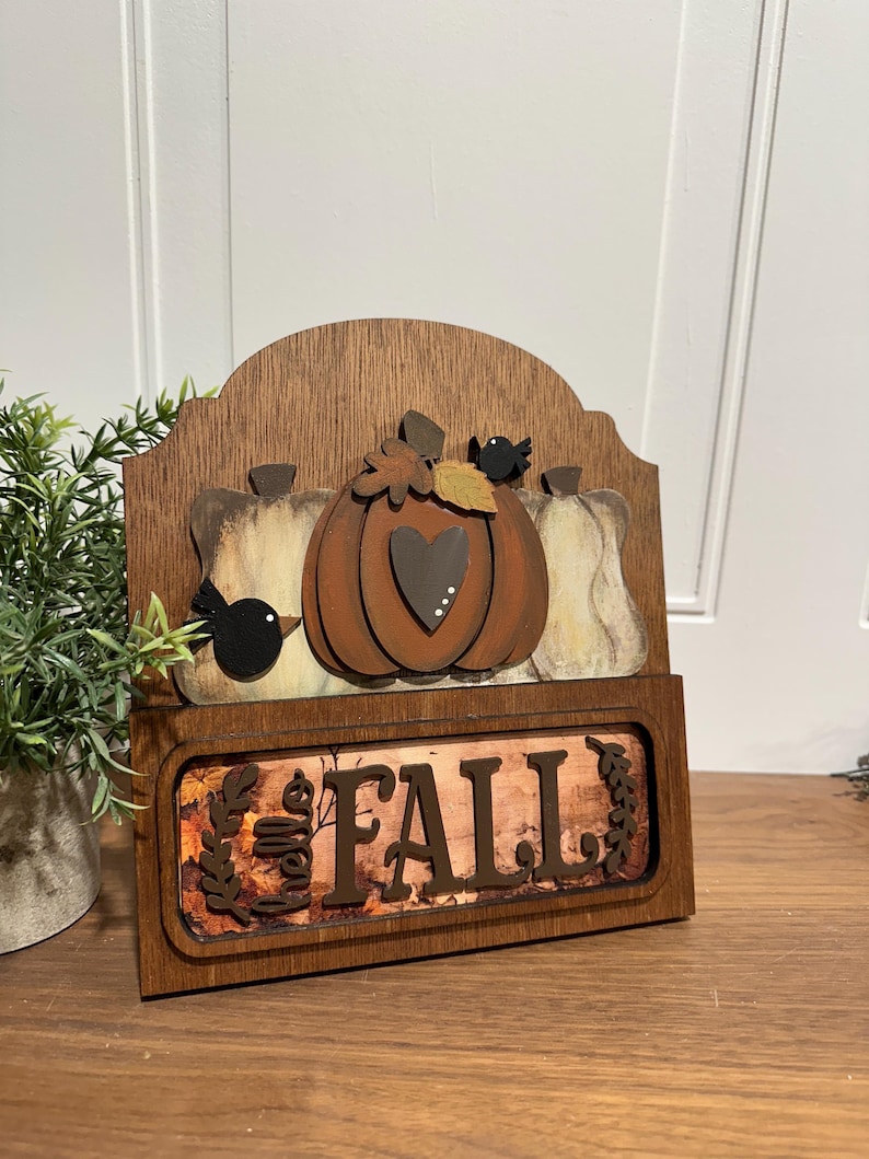 May include: Wooden sign with a autumnal theme. It features a brown pumpkin with a heart, two white pumpkins, and two black birds. The sign also has a brown background with the words "Hello Fall" in white letters.