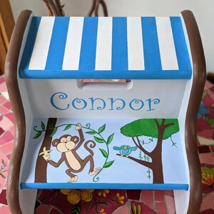 May include: A blue and white striped wooden step stool with a jungle theme. The stool is painted with a monkey, a bird, and a tree. The name "Connor" is painted on the top step.
