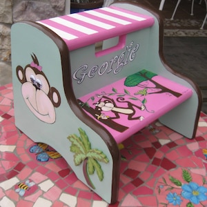 May include: A light blue and pink wooden step stool with a monkey and palm tree design. The stool has a pink top with white stripes and a cutout handle. The name "Georgie" is painted on the front of the stool.