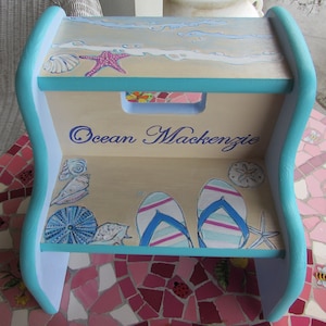 May include: A teal and light blue wooden step stool with a beach theme. The top step has a painted scene of waves, a starfish, and seashells. The bottom step has a painted scene of flip flops, a sea urchin, seashells, and a starfish. The stool is personalized with the name "Ocean Mackenzie".