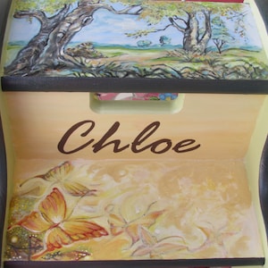 May include: A wooden step stool with a painted scene of trees and butterflies. The stool is painted in shades of yellow, brown, and green. The name "Chloe" is painted on the front of the stool.