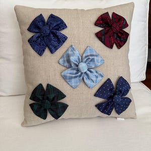 May include: A beige square pillow adorned with five decorative bow accents in various patterns and colors. The bows include navy blue paisley, red plaid, light blue plaid, dark green plaid, and navy blue with a small white pattern.