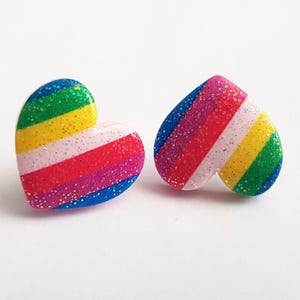 Op de afbeelding: Twee hartvormige oorknopjes met een regenboog gestreept ontwerp en glitteraccenten. De oorbellen hebben een levendig kleurenpalet van blauw, groen, geel, roze en rood. De oorbellen staan tegen een witte achtergrond.