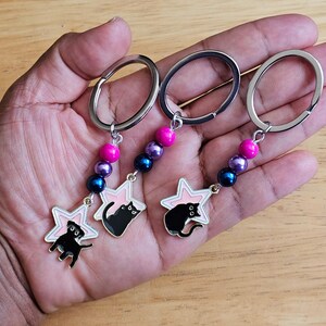 May include: Three silver-toned metal keychains, each featuring a star-shaped charm with a black cat silhouette. The charms are attached to a silver ring by a chain of three beads in bright pink, metallic purple, and dark blue colors.