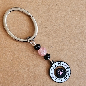 May include: A silver key ring with black and pink beads and a black charm with the text "Team Toe Beans".