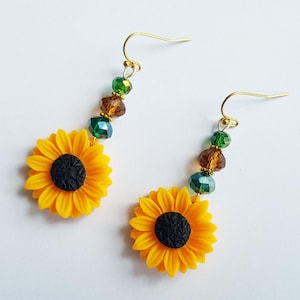 May include: A pair of drop earrings featuring yellow sunflower charms with black centres. Each earring hangs from a gold-tone hook and includes a vertical arrangement of three faceted beads in alternating shades of green and brown.