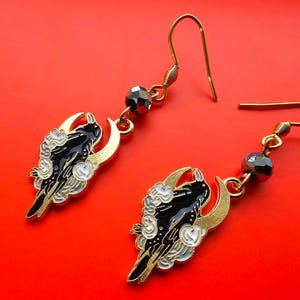 Puede incluir: Un par de pendientes de oro con diseños de cuervo negro y luna. Cada pendiente presenta un cuervo negro posado en una luna creciente con nubes blancas. Los pendientes tienen detalles de cristal negro.