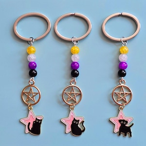 Può includere: Tre portachiavi con un anello argentato collegato a una fila verticale di quattro perline gialle, bianche, viola e nere. Sotto le perline pendono un ciondolo a forma di pentagramma dorato e una stella rosa con la sagoma di un gatto o cane.