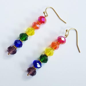 Puede incluir: Un par de pendientes de cuentas coloridas. Cada pendiente presenta un degradado de cuentas facetadas en rojo, naranja, amarillo, verde, azul y morado. Las cuentas están ensartadas en ganchos dorados, creando un efecto arcoíris.
