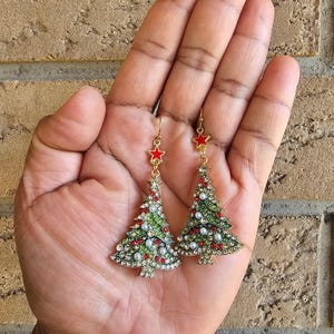 Puede incluir: Un par de pendientes de árbol de Navidad de tono dorado con estrellas rojas, esmalte verde, perlas blancas y pedrería transparente.