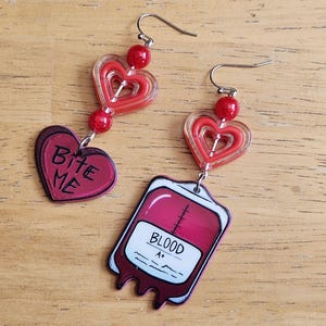Peut inclure: Une paire de boucles d'oreilles fantaisie. Une boucle d'oreille représente un cœur rouge avec le texte "Bite Me". L'autre boucle d'oreille a un design de poche de sang avec le texte "BLOOD A+". Les deux ont des perles rouges et des crochets d'oreilles argentés.