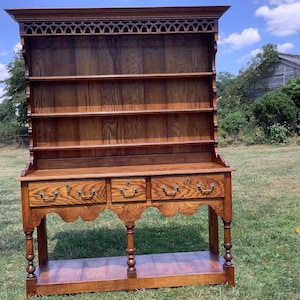 May include: An oak dresser with a rich, medium-brown stain. It has open shelves, three drawers with brass handles, and decorative fretwork. The dresser is supported by turned legs and is positioned on a grassy area, likely a garden.