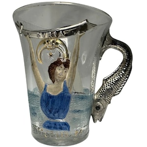 May include: A clear glass mug with a silver fish handle. The mug features a painted image of a woman holding a banner that reads "SYRIA" above a crescent moon and star. The bottom of the mug reads "PITTSBURG PA".