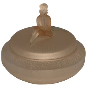 May include: A frosted glass trinket box with a lid featuring a sculpted figurine of a woman sitting on the lid. The box is a pale pink color.