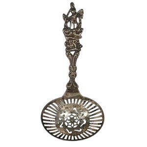 May include: Antique silver-toned sugar spoon with a decorative handle featuring a figure and ornate floral designs. The spoon has a slotted, oval bowl with a floral pattern in the center. A vintage kitchen utensil.
