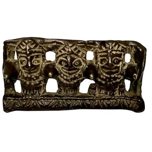 May include: A bronze decorative piece with three stylized faces, each with a crown and a beaded necklace. The piece has a textured surface and a decorative border.