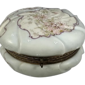May include: A white porcelain trinket box with a hinged lid. The lid is decorated with a floral design in shades of pink, white, and green. The box has a gold-toned metal hinge.