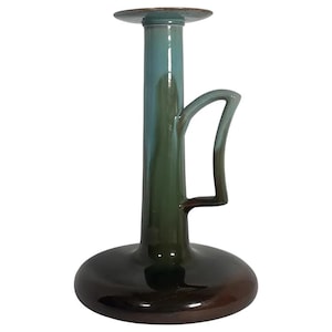May include: A ceramic candlestick holder with a teal, green, and brown glaze. The candlestick has a wide base, a tall stem, and a handle. The top is a flat disc. The overall design is modern and artistic.