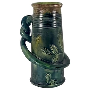 May include: A green and brown ceramic pitcher with a decorative handle. The pitcher features embossed designs of a sun, leaves, a bird, and a duck. The handle is a twisted rope design. The pitcher is approximately 8 inches tall.