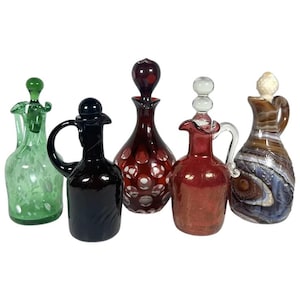 May include: A collection of five decorative glass bottles in various colors. The bottles include green, black, red, and brown, with unique shapes and stoppers. Some bottles have etched designs or handles, showcasing a vintage aesthetic.