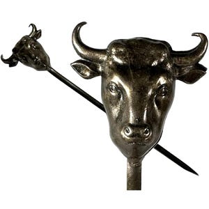 May include: Two silver-toned metal objects with bull head designs. One is a bull head on a stick, and the other is a bull head with a long, pointed rod through it. The objects are likely decorative or functional.
