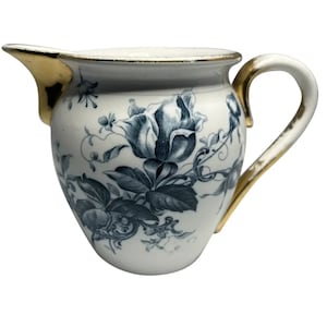 May include: A white ceramic creamer with a gold rim and handle. The creamer is decorated with a blue and white floral pattern.