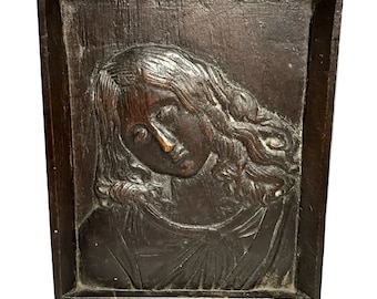 May include: A dark wood framed relief carving of a woman's head with long flowing hair. The carving is in a simple frame and the woman's face is in profile.