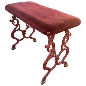 May include: A burgundy bench with a rectangular, cushioned seat. The bench has ornate, red metal legs with a scrolled design. The bench is a decorative piece of furniture.