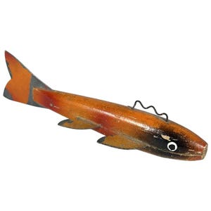 May include: Wooden fish figurine with orange, red, and black paint. The fish has a wire loop for hanging. The tail and fins are orange, the body is a gradient of orange and red, and the head is black with a white eye. A decorative item.