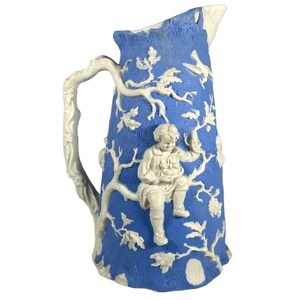 May include: A blue and white ceramic pitcher with a raised design of a child in a tree with birds. The pitcher has a handle and a spout.